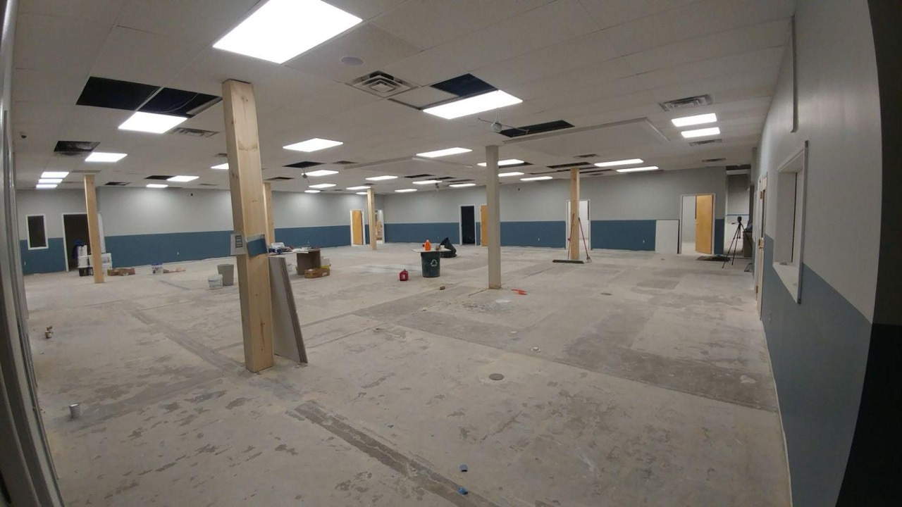 Large commercial interior under construction with floor preparation, ceiling work, columns, lighting, and painted walls