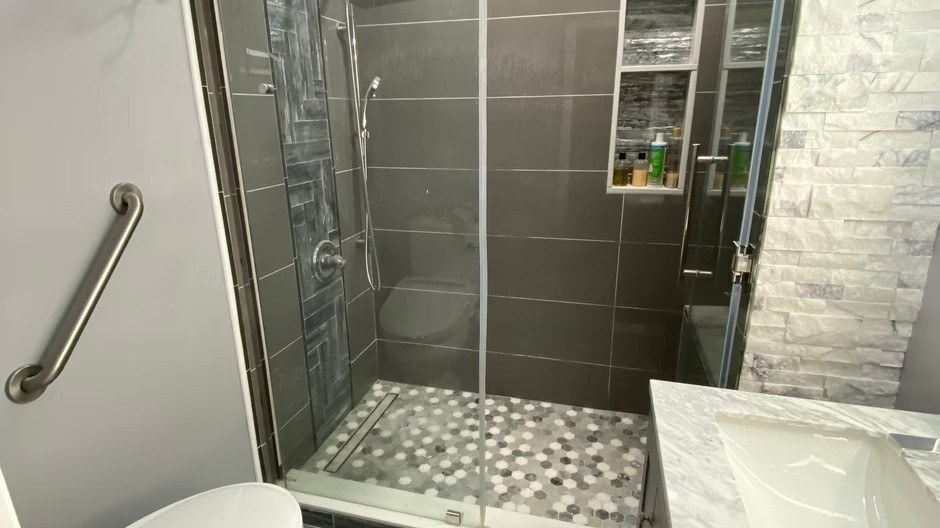 Gray tiled shower with hex-mosaic floor, recessed shelves, glass enclosure, and handheld fixture